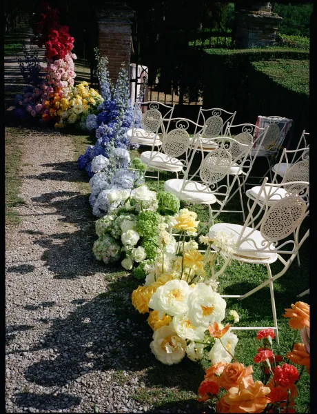 Ceremony aisle design with outdoor ceremony seating, lined by ground florals along a gravel path between garden hedges and an iron gate.