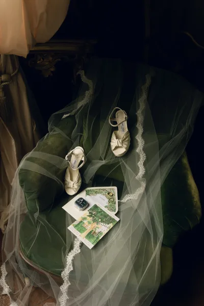 Bridal shoes, ivory bridal heels paired with a soft wedding veil, arranged on a velvet chair with gentle drapery in the background.