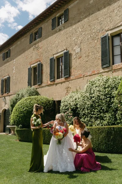 Bride and bridesmaids share a candid bridal party moment before a stone villa, the bride holding a colorful bridal bouquet.