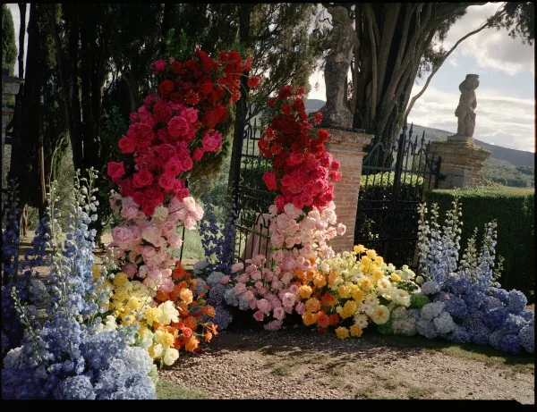 Ceremony floral arch shaping a garden ceremony backdrop, lush with roses, set before stone pillars and a wrought iron gate amid trees and hedges.