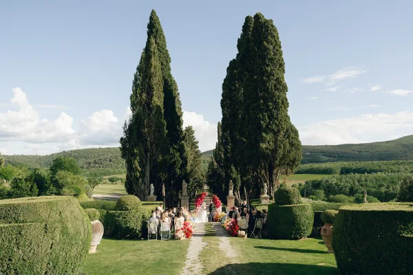 Garden ceremony setup featuring elegant outdoor ceremony decor with colorful floral installations along a gravel aisle framed by cypress trees.