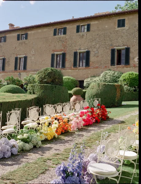 Ceremony aisle design with outdoor ceremony decor, lush florals lining a gravel path before a stately stone building and clipped garden hedges.