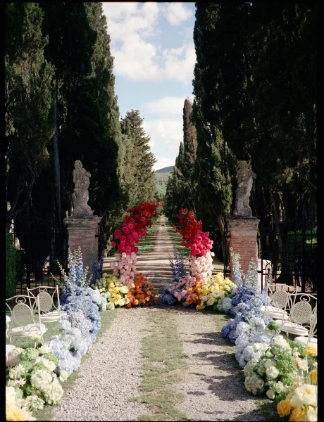 Ceremony aisle design showcasing outdoor ceremony decor with a floral arch at the entry along a gravel path beneath tall trees and distant hills