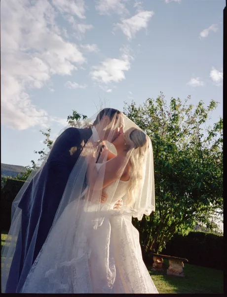 Wedding kiss portrait as a bride and groom kiss beneath a flowing bridal veil, with blue sky and garden trees soft in the background.