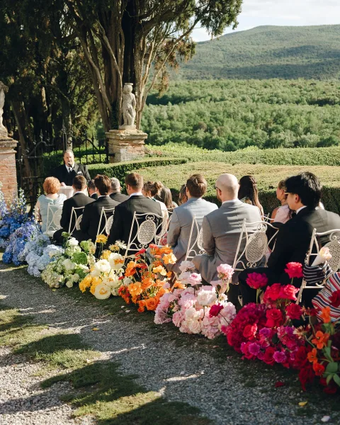 Ceremony aisle flowers anchor outdoor ceremony decor along a gravel path, flanked by white metal chairs, with a stone gate and mountains beyond.