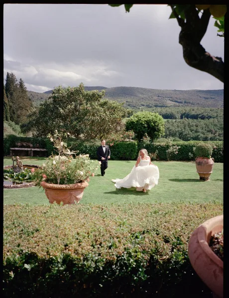 Couple portrait of a bride and groom in garden, standing on a manicured lawn by a garden fountain, framed by hedges beneath a cloudy sky.