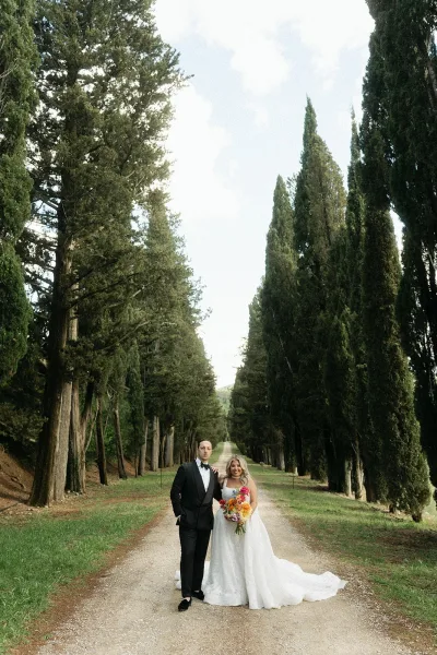 Couple portrait of a bride and groom on path under a cypress tree tunnel, the bride holding a bridal bouquet as they stand on a gravel lane.