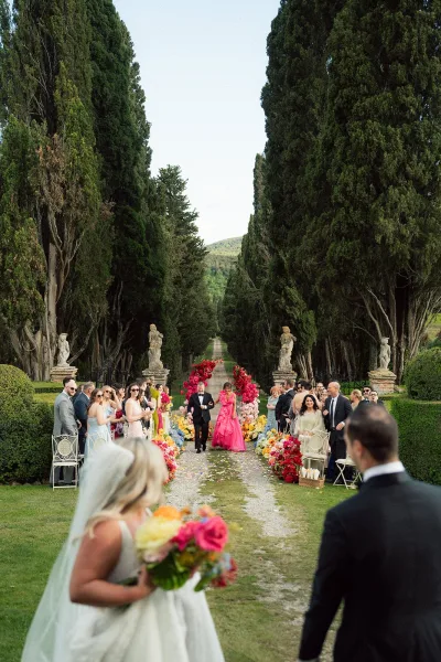 Ceremony aisle design anchored by a floral arch, showcasing garden ceremony decor framed by cypress trees and stone statues.