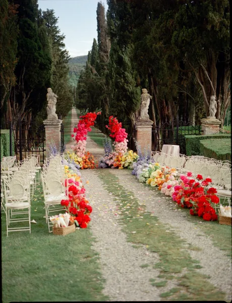 Ceremony setup with refined outdoor ceremony decor centered on a floral arch before a wrought iron gate amid cypress trees and hills.