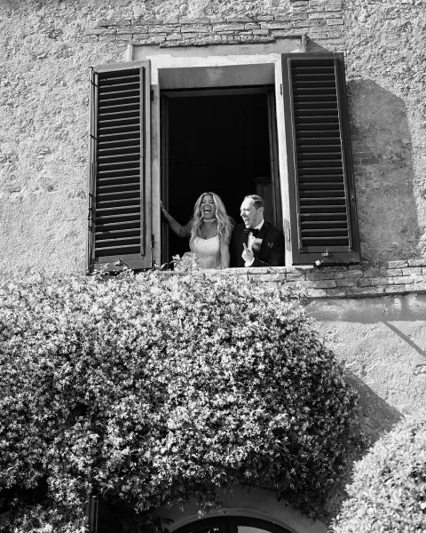 Couple portrait of a bride and groom at a window, groom in a black tuxedo, framed by shuttered panes on a vine-covered stone wall.