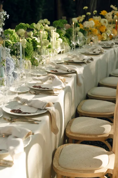 Reception tablescape styled for garden reception decor, lined with taper candles and set outdoors amid lush garden greenery and trees.