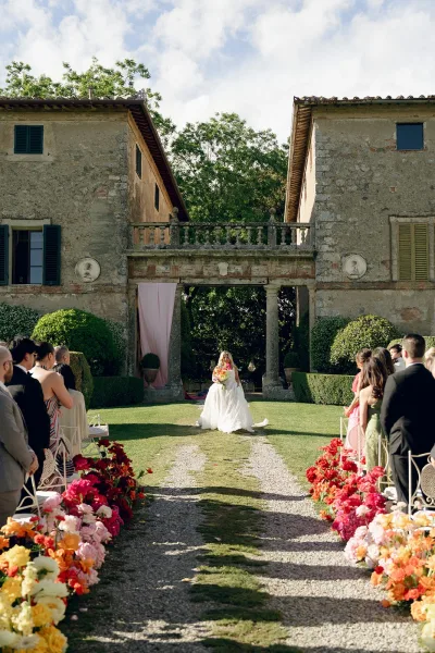 Ceremony processional with a bride walking down aisle past bright aisle flowers toward a stone villa on a gravel path.