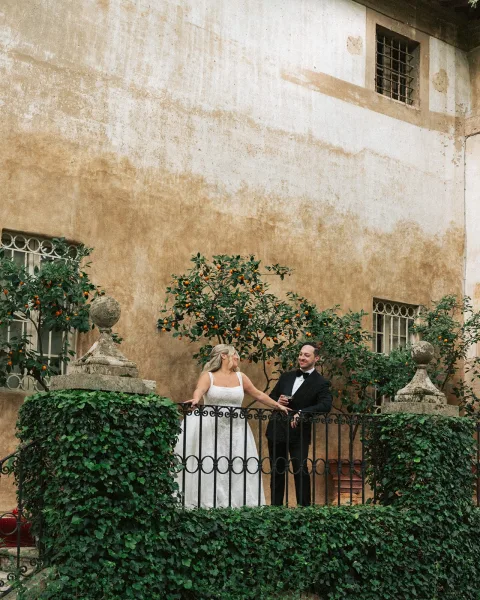 Couple portrait of a bride and groom on balcony, resting by a stone balustrade in a rustic stucco courtyard with garden greenery.