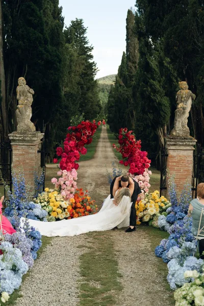 wedding kiss moment as newlyweds share an aisle kiss beneath a floral arch, framed by cypress trees and stone pillars on a gravel path.