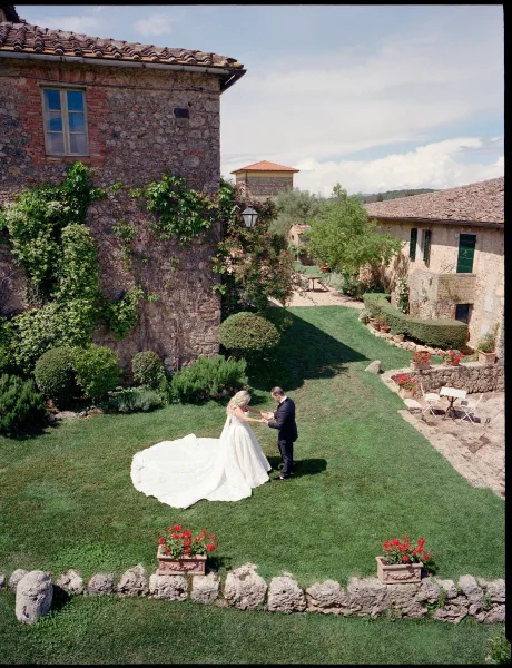 Couple portrait of a bride and groom holding hands on a courtyard lawn, her bridal gown set against ivy-clad stone buildings under blue sky.