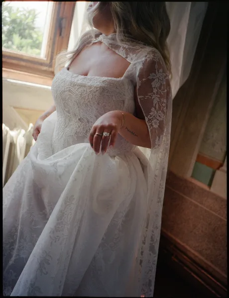 Wedding dress close-up of a lace wedding dress with a lace capelet, beside a window with sheer curtains.