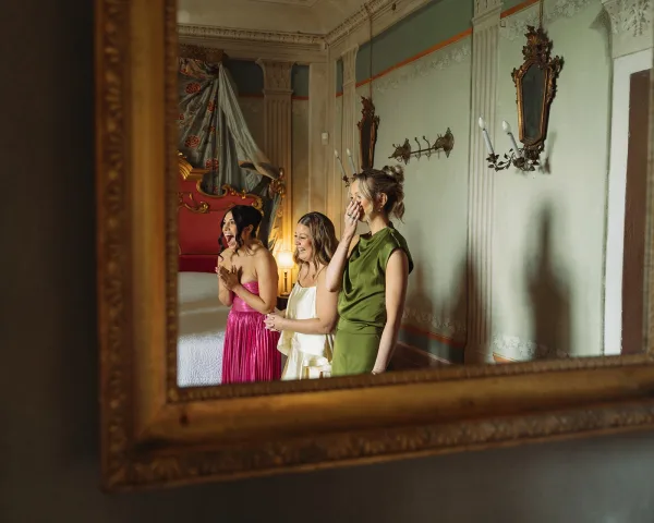 Bridesmaids first look during a getting ready moment, clapping in satin dresses in a bridal suite with a canopy bed and mirrors.