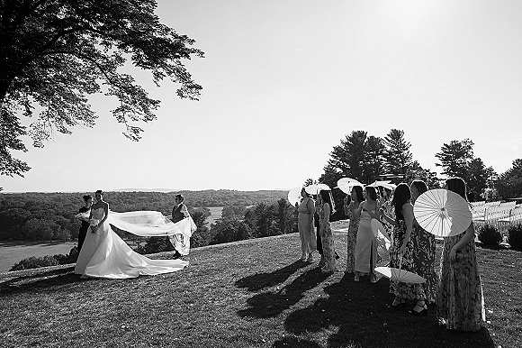 Bridal party portrait with bride with bridesmaids, long veil and bouquet, group holding white parasols on a grassy hill by trees