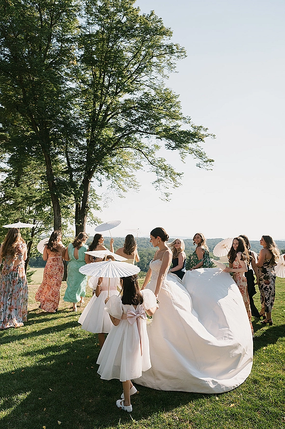 Bridal party portrait with bride with bridesmaids holding parasols, bouquet in hand, on a grassy lawn under a large tree with rolling hills.