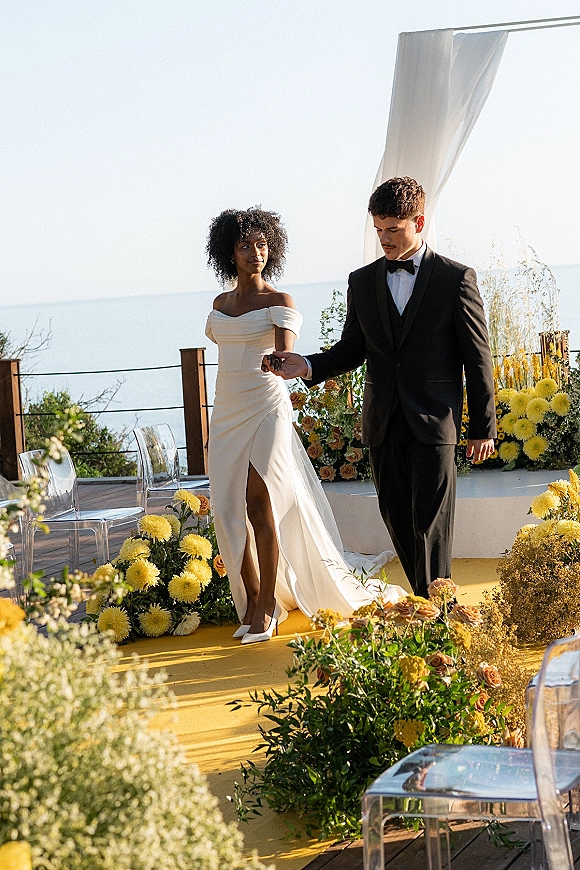 Ceremony recessional as bride and groom walking aisle hand in hand on a yellow runner, ocean view behind; her off-shoulder gown train flows