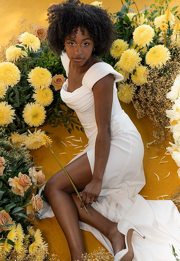 Bridal portrait of a bride in an off shoulder wedding dress sitting amid yellow chrysanthemums and peach roses on a mustard yellow backdrop