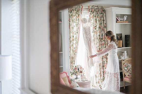 Wedding dress hanging by a window with lace accents, with bouquet and shoes nearby in a romantic bridal room with floral curtains