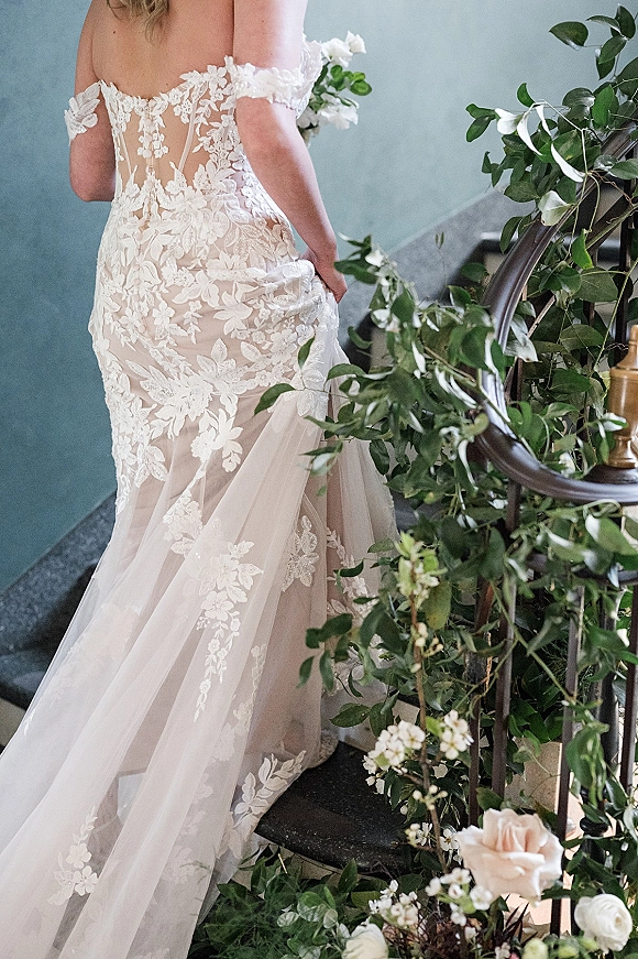 Wedding dress with lace wedding dress details, off-the-shoulder straps and tulle train flowing up a staircase with greenery garland on railing