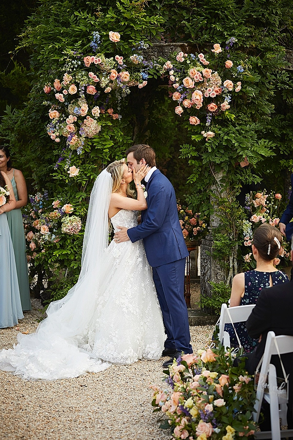 Ceremony kiss as bride in lace gown and long veil kisses groom in blue suit beneath stone arch with peach and blue florals