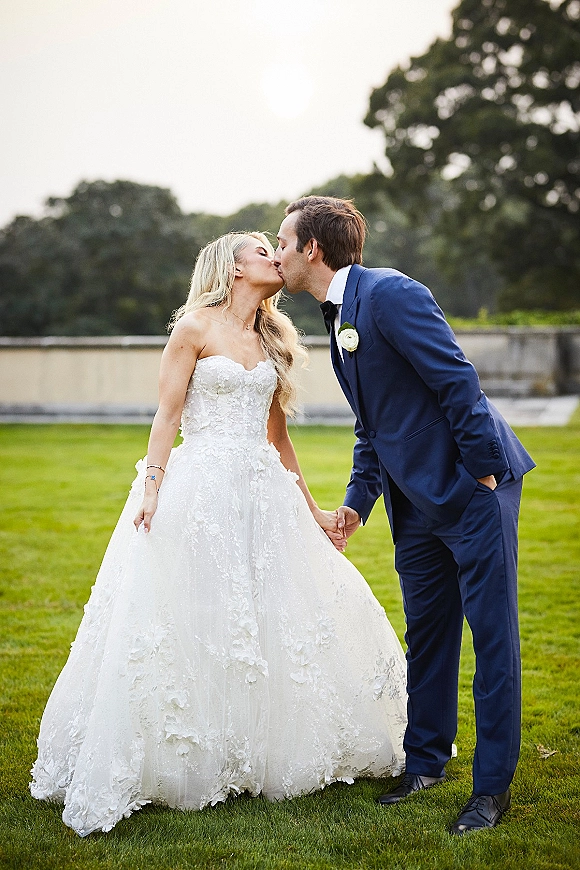Wedding kiss portrait of bride and groom kissing, her strapless lace gown and his blue suit on a green lawn by a stone wall
