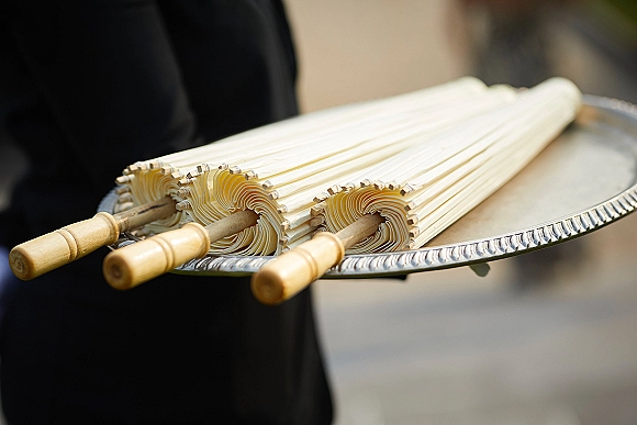 Wedding parasols with bamboo handles arranged on a silver tray, ready for guests along an outdoor walkway beside black-clad attendants