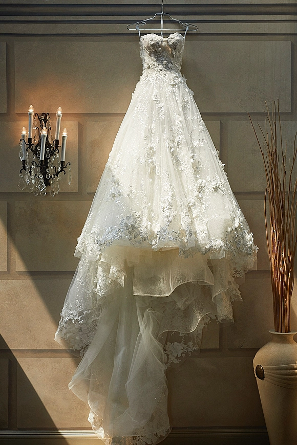 Wedding dress hanging on a dress hanger, strapless lace appliques and layered tulle skirt against a paneled wall with warm sconce light