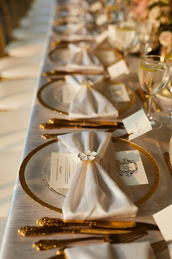 Reception tablescape with gold rim charger plates, white napkins and floral centerpiece on a long banquet table in warm ambient light