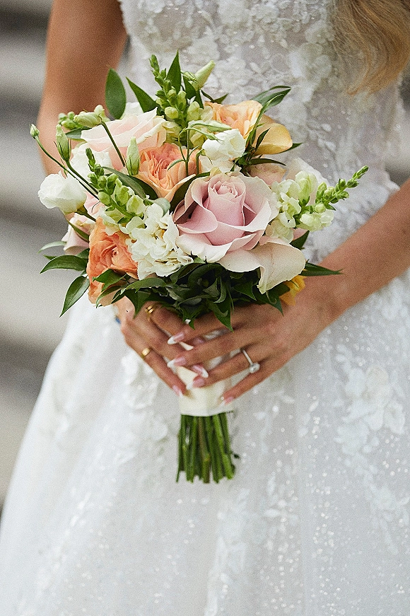 Bridal bouquet of peach and blush roses with greenery, held against a lace wedding dress with ringed hand and soft neutral backdrop