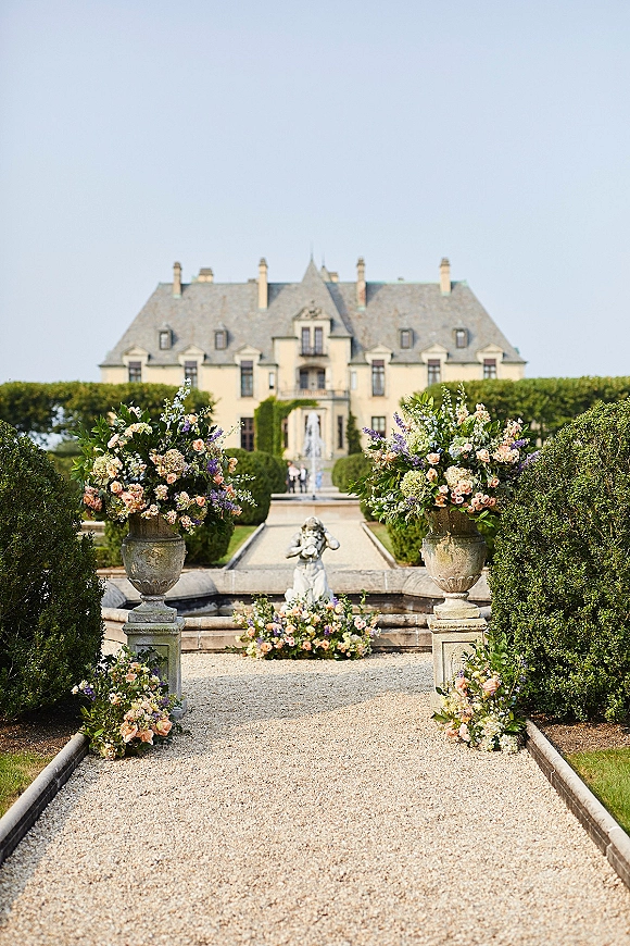 Wedding ceremony aisle lined with outdoor wedding aisle decor, stone urns and pastel pink-purple florals along a gravel path by a chateau and fountain