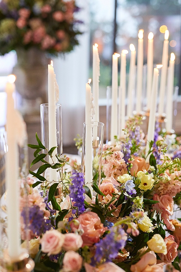 Wedding tablescape with a taper candle centerpiece, glass cylinder candles, and rose greenery florals beside a bright reception window