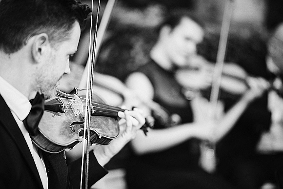 Wedding musicians in a string quartet playing violins in tuxedos, bows poised on strings with other players and blurred crowd behind