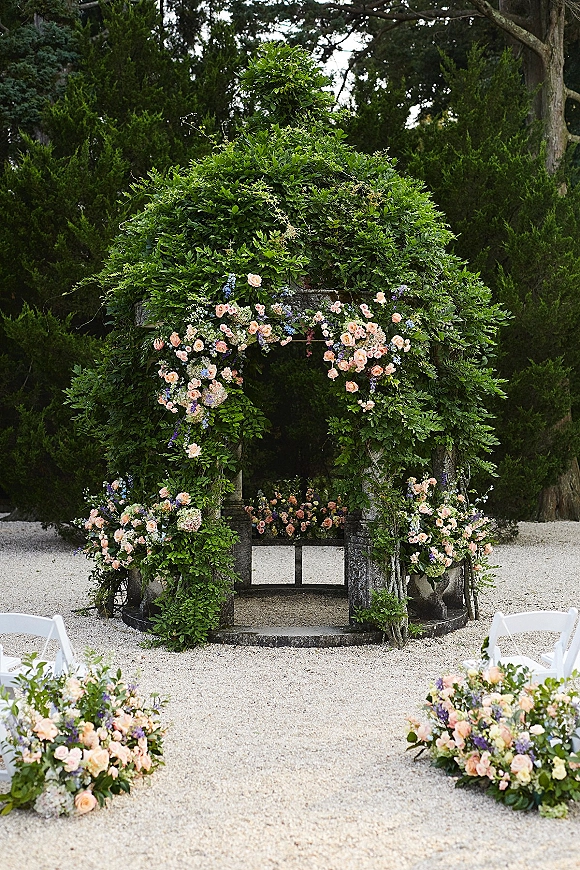 Wedding ceremony arch draped in greenery wedding arch vines with roses and blue flowers, flanked by white chairs on gravel in a tree-filled garden