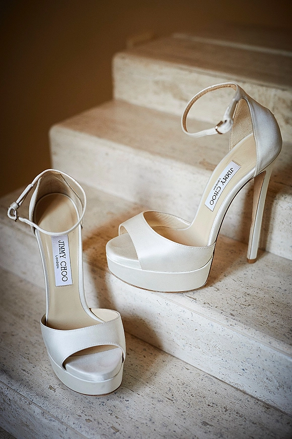 Wedding shoes in ivory bridal heels with ankle straps resting on a stone staircase against an indoor wall for a getting-ready detail shot