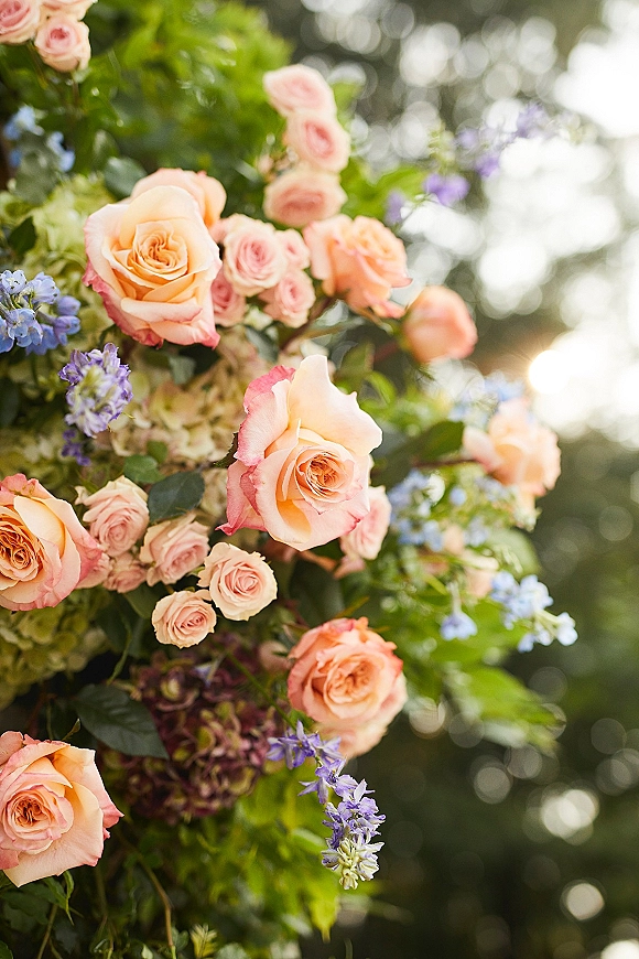 Wedding floral arrangement with peach rose wedding flowers, blue blooms and hydrangea amid greenery, softly lit by sun and blurred trees