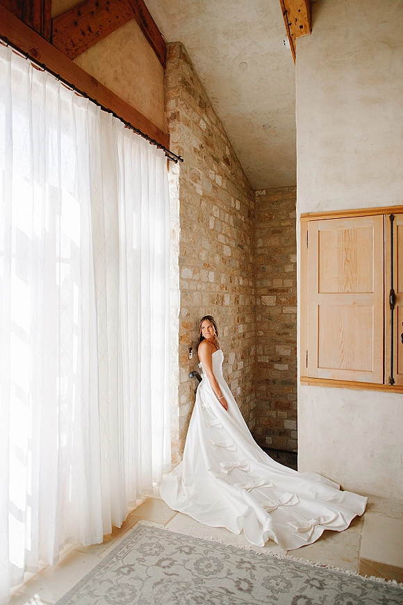 Bridal portrait of a bride in a strapless wedding dress with a long bow-trimmed train, posing by window light in a stone room.