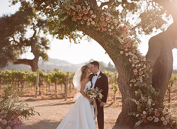 Wedding kiss as bride and groom embrace beneath a rose floral arch in a sunlit vineyard, her veil and bouquet glowing at golden hour