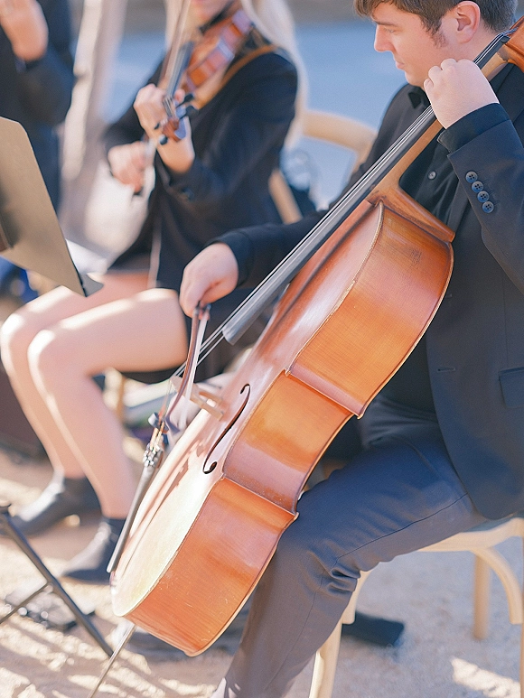 Wedding musicians in black suits playing cello and violin, bows moving over strings beside a sheet music stand on outdoor pavement in soft daylight