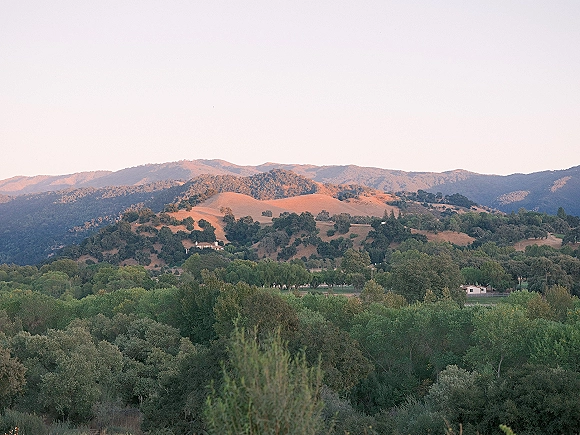 Mountain landscape with rolling hills landscape at golden hour, showing a tree-covered valley, scattered houses, and distant peaks under a soft sky
