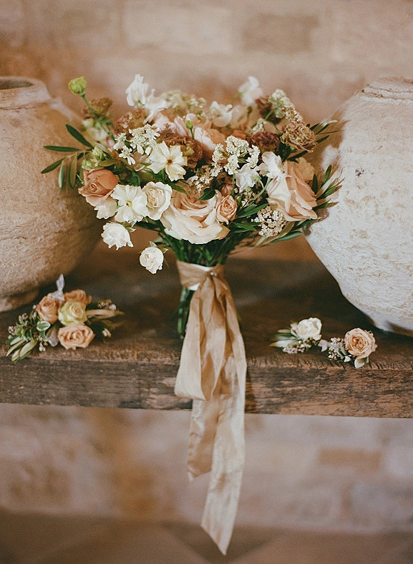 Bridal bouquet of blush roses, white flowers, and greenery with a ribbon wrap on a wooden bench beside a matching boutonniere
