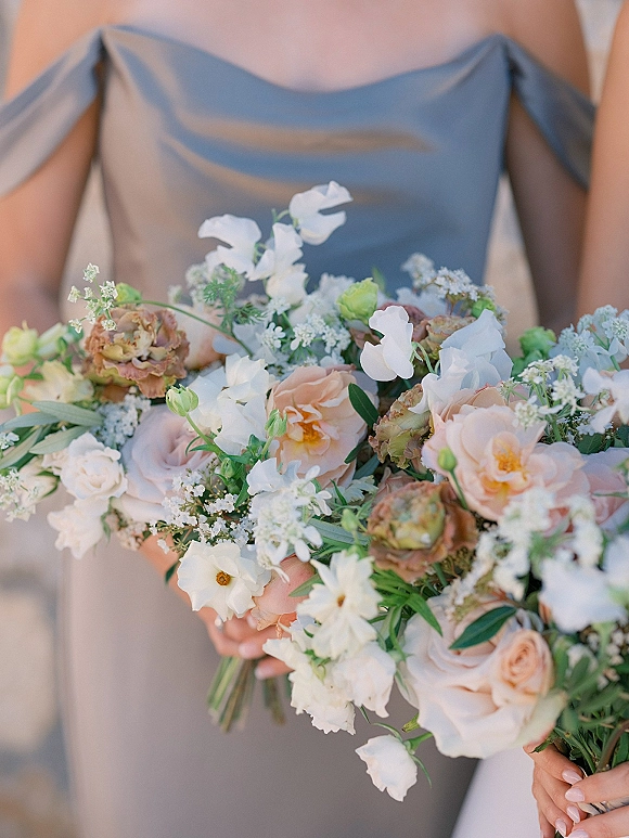 Bridesmaid bouquet of blush and white flowers with peach roses, sweet peas, baby’s breath, and greenery held against a neutral backdrop
