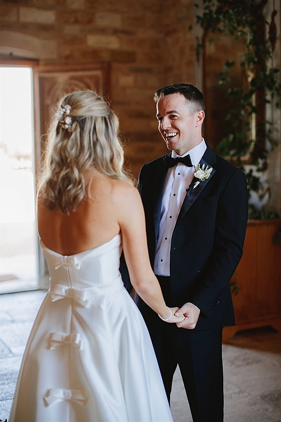 First look moment as bride in strapless satin wedding dress holds hands with tuxedo groom by wooden walls and potted greenery indoors