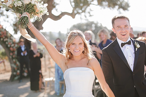 Wedding recessional as bride raises bouquet with ribbon streamers while newlyweds walk the outdoor aisle past cheering guests and arch
