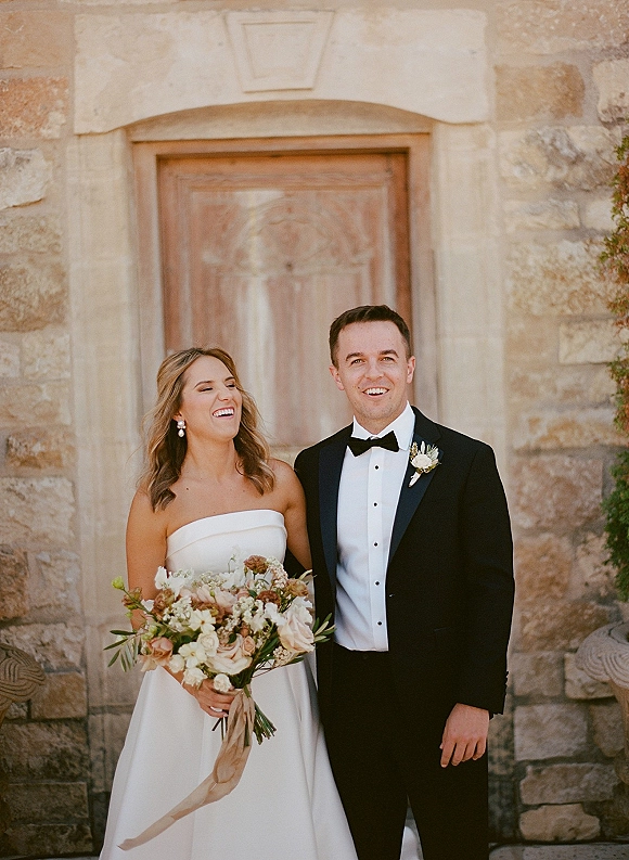 Couple portrait of bride in a strapless wedding dress holding a bouquet, beside groom in black tuxedo by a stone wall and wooden door