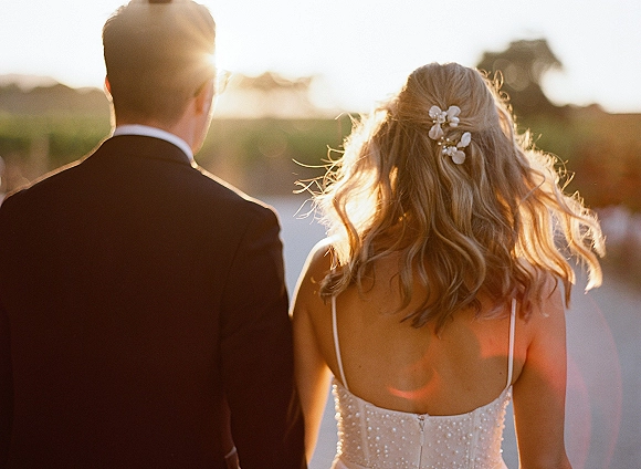 Couple portrait of bride and groom from behind, walking by the water at sunset, her pearl beaded bodice and loose waves glowing
