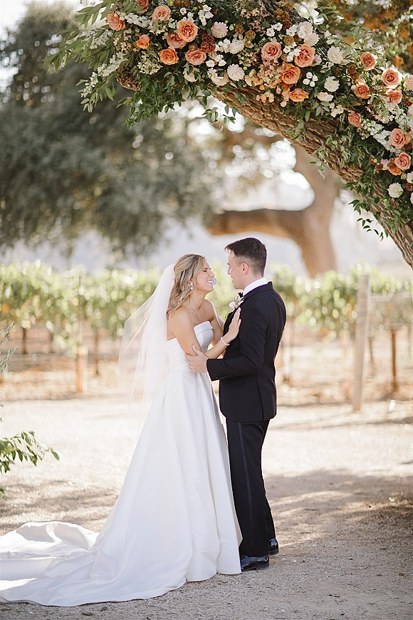 Couple portrait of bride and groom embrace under a rose and greenery floral arch, cathedral veil and tuxedo in sunlit vineyard rows